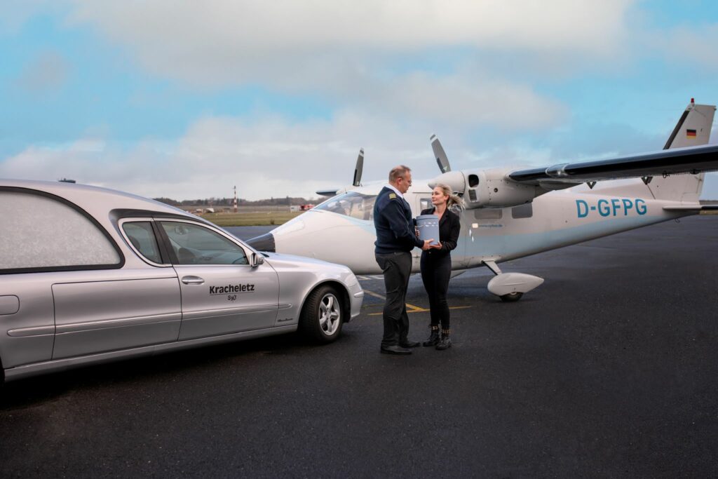 Übergabe einer Urne auf einem Flugplatz. Auf dem Flugplatz steht ein Bestattungswagen und ein Flugzeug vom Typ Cirrus SR20.